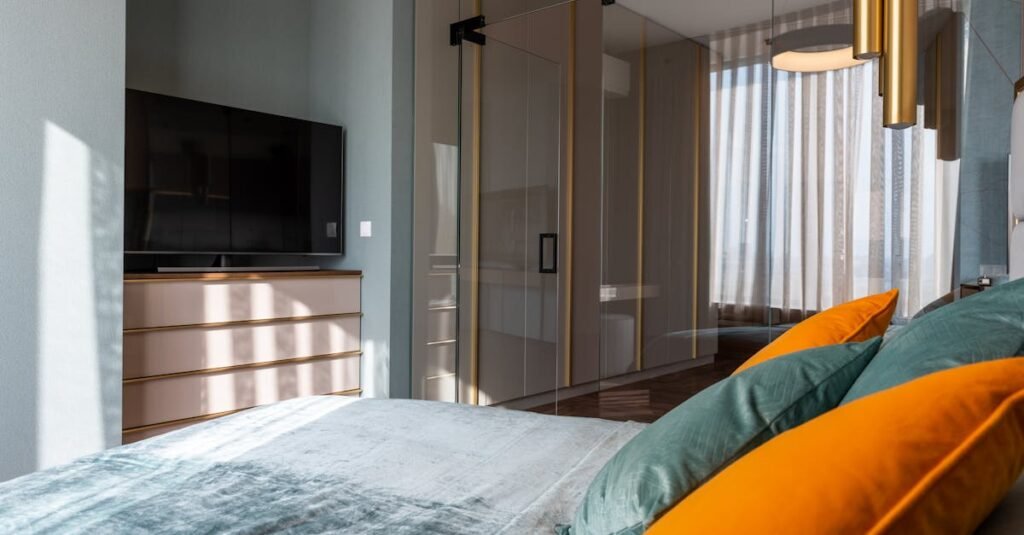 Interior of modern bedroom with colorful pillows on bed placed against TV with drawer near glass door in contemporary apartment